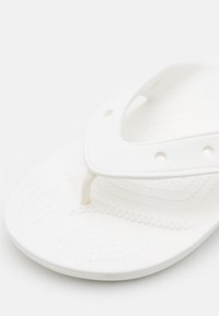 Valkoiset flip-flopit, joissa on teksturoitu pohja, pehmeät kumihihnat ja minimalistinen muotoilu. Hihnan lähellä on pieni logoyksityiskohta.