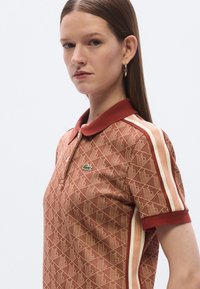 Póló ing vörösesbarna színben, geometriai mintával, bordázott gallérral, gombolós szegéllyel és krémszínű, valamint bordó hangsúlyos csíkokkal. A mellkason zöld logó található.