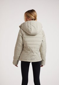 Lys grå dunjakke med hætte, prydet med et quiltet mønster og trekantede detaljer på ryggen. Sorte leggings ses nedenunder.