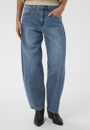 Donna che indossa jeans blu larghi con tasche, top beige, bracciali dorati e stivaletti neri con tacco su sfondo bianco.