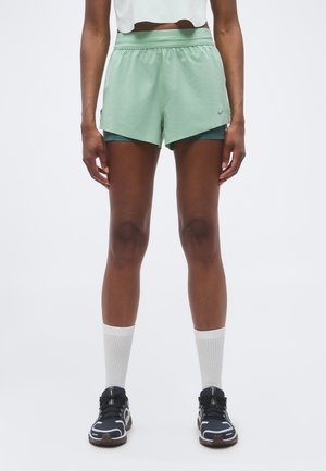 Personne portant un short de sport vert clair avec une couche de compression intérieure, des chaussettes blanches montantes, des chaussures de course noires et blanches, debout avec les bras détendus.