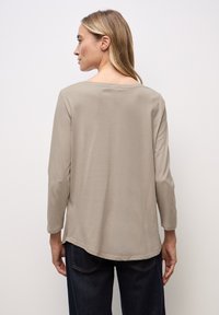 Beige Langarmhemd mit weitem Ausschnitt, weichem Material und geschwungener Saum, kombiniert mit dunkelblauen Jeans.