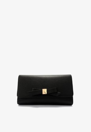 Bolso de mano de cuero negro con textura, con un lazo plano como detalle y un pequeño broche cuadrado de tono dorado en el centro, sobre un fondo blanco.