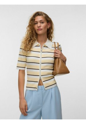 Kvinde med krøllet hår iført en lynlåst, stribet kortærmet top, lyseblå bukser og bærende en beige trekantet håndtaske på skulderen.