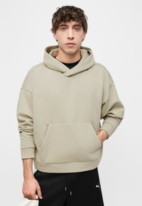 Νεαρός άνδρας που φοράει μπεζ φούτερ με κουκούλα και τσέπη καγκουρό, καθώς και μαύρη φόρμα Puma, στέκεται μπροστά σε απλό λευκό φόντο.