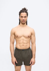 Homme torse nu avec des dreadlocks portant un short vert olive foncé, debout devant un fond uni gris clair, faisant face à l'avant.