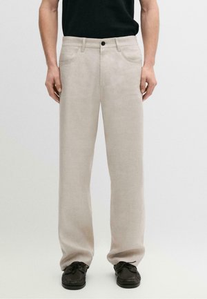 Homme portant un pantalon beige droit avec des poches avant, une boucle de ceinture noire et des chaussures décontractées noires sur un fond clair uni.