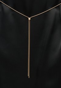 Λεπτό κολιέ με χρυσή αλυσίδα και στρογγυλό κούμπωμα στο κέντρο, με δύο κρεμαστές αλυσίδες πάνω σε μαύρο υφασμάτινο φόντο.