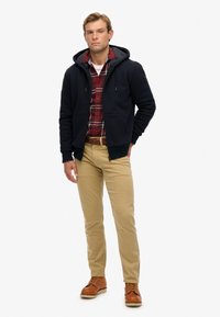 Mørkeblå zip-up hoodie med grå inderside, båret over en rød- og sortternet skjorte, parret med beige bukser og brune støvler.