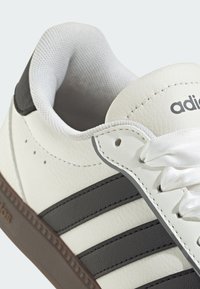 White leather sneaker with three dark gray stripes, padded collar, and white laces. Features textured surfaces and an embossed logo.