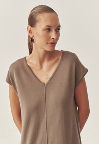 Brauner V-Ausschnitt-Pullover mit kurzen Ärmeln, aus weichem Stoff. Auffällige zentrale Nahtdetails. Getragen mit lila dreieckigen Ohrringen.