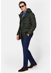 Zielona kurtka ocieplana z kapturem i przednimi kieszeniami, w połączeniu z ciemnoniebieskimi spodniami i brązowymi zamszowymi butami. Dodatki to czarne okulary przeciwsłoneczne.