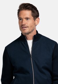 Donkerblauwe zip-up jas van zachte stof met een gestructureerde breiwerk op de mouwen en een hoge kraag, gecombineerd met een witte binnen tshirt.