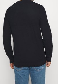 Svart stickad tröja med ett texturerat mönster, långa ärmar och ribbade mudd. Bärs tillsammans med ljusblå denimjeans.