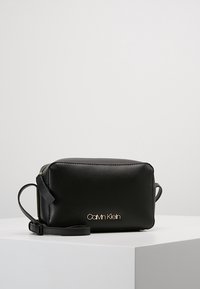 Černá kožená crossbody taška s obdélníkovým tvarem, zlatým logem "Calvin Klein" a odnímatelným popruhem. Hladká textura, minimalistický design.