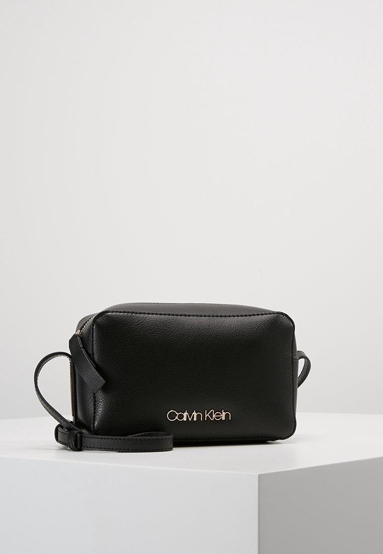 Černá kožená crossbody taška s obdélníkovým tvarem, zlatým logem "Calvin Klein" a odnímatelným popruhem. Hladká textura, minimalistický design.