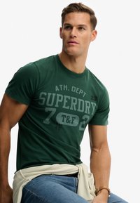 T-shirt en coton vert avec col rond, arborant un grand texte gris "SUPERDRY" et des chiffres. Porté avec une montre beige et un jean bleu clair.