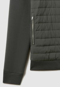 Veste grise foncée avec une section en tissu matelassé, dotée d'une fermeture éclair argentée et de poignets côtelés. Texture lisse sur les manches et le corps.