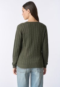 Donna con lunghi capelli castani che indossa un maglione verde oliva a trecce e jeans azzurri, di spalle contro uno sfondo bianco semplice.