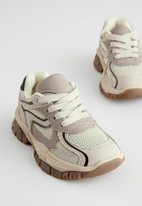 Lys beige atletiske sneakers med mesh materiale, grå accenter, tykke gummisåler og traditionelt snøresystem. Tekstureret finish overalt.