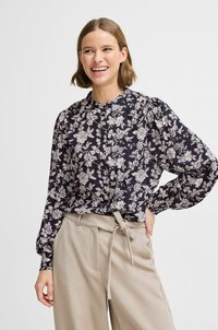 Blusa a maniche lunghe nera con motivo floreale bianco, caratterizzata da un colletto alto e una chiusura frontale a bottoni, abbinata a pantaloni beige a vita alta.