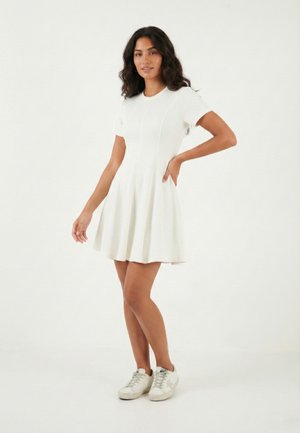 Kobieta stojąca z ręką na biodrze, ubrana w krótki biały rękaw sukienki i białe trampki na jednolitym białym tle.