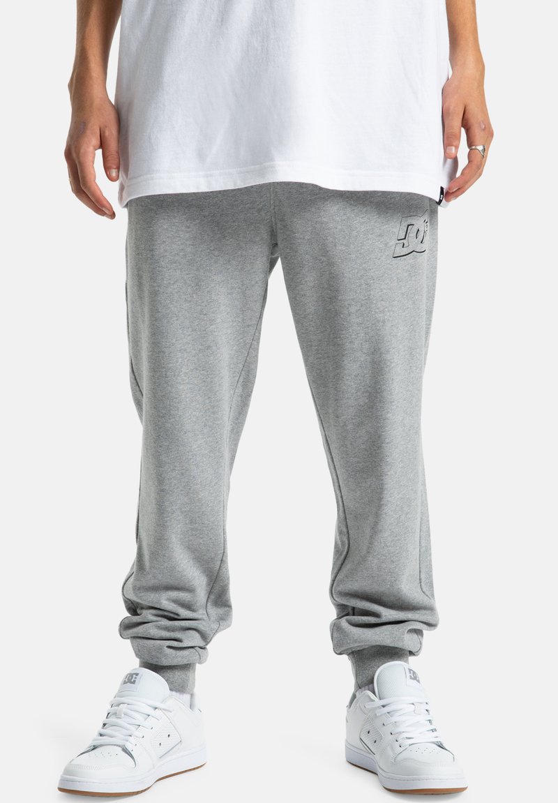 Graue Sweatpants aus weichem Material mit lockerer Passform, am Knöchel umschlagen, mit einem DC-Logo auf dem linken Oberschenkel und kombiniert mit weißen Sneakers.