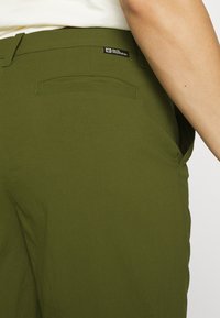 Olivgröna byxor med en slät yta, som har en bakficka och en liten märkesetikett på midjebandet. Minimalistisk design.