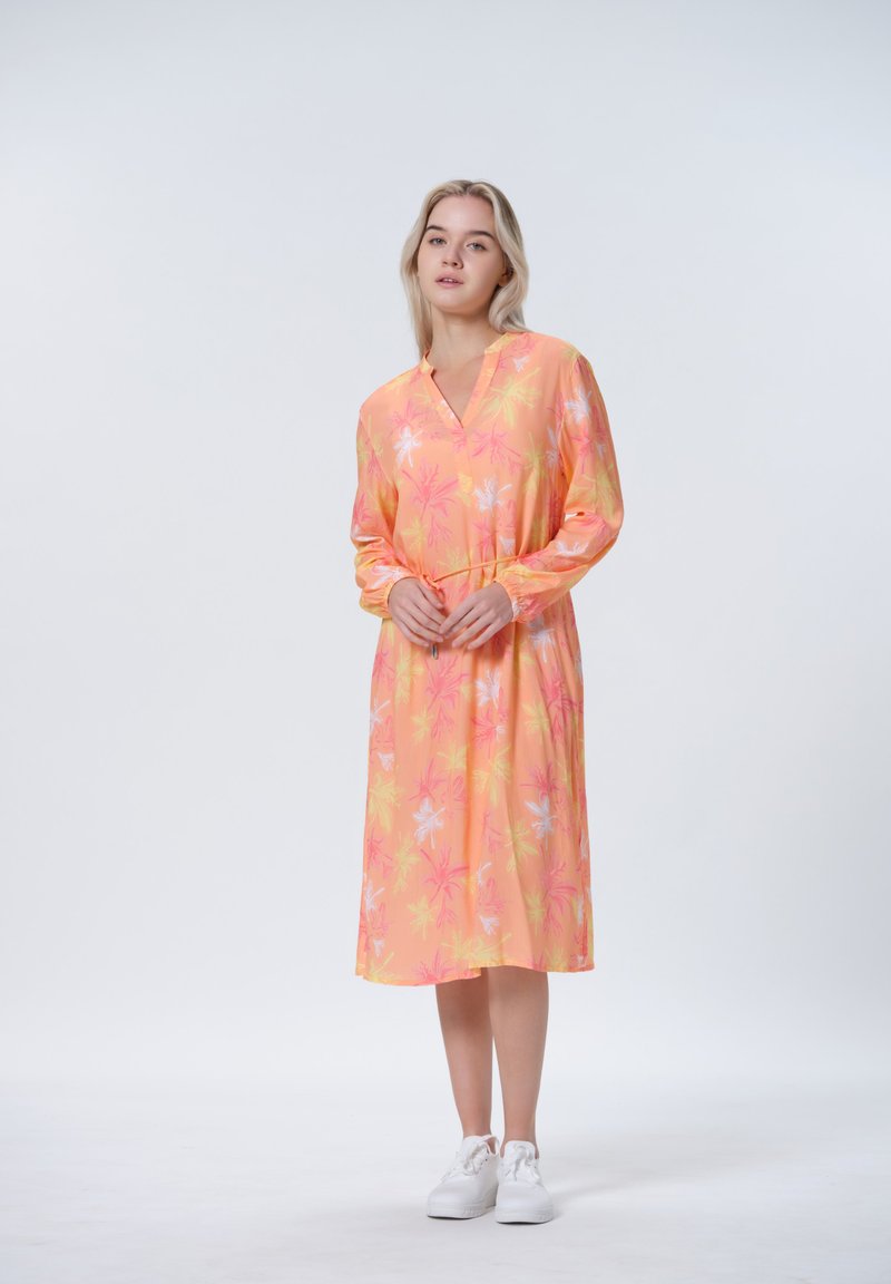 Langärmliges Kleid in sanftem Gelb und Rosa mit Palmenblattmuster. Hat einen V-Ausschnitt und eine taillierte Taille, kombiniert mit weißen Sneakers.
