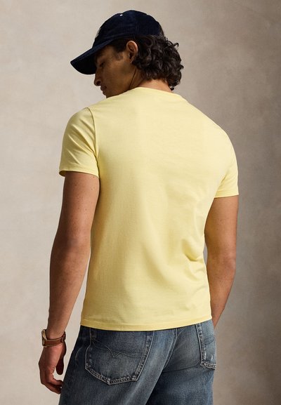 Moški, ki nosi bledo rumeno majico s kratkimi rokavi, modre jeansa, mornarsko modro kapo in rjavo uro, obrnjen z zadnje strani na enobarvnem ozadju.