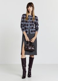 Grauer Rock mit seitlichem Schlitz, gemusterte Strickjacke in Blau und Burgunderrot, braune Lederhandtasche und glänzende kniehohe Stiefel.