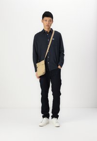 Camicia blu navy con bottoni, jeans scuri, borsa a tracolla beige, sneakers bianche. L'outfit presenta una vestibilità comoda e elementi di design minimalisti.
