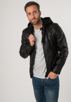 Schwarze Lederjacke mit Kapuze, ausgestattet mit einem Reißverschluss und gerippten Akzenten. Getragen über einem hellgrauen Shirt mit blauen Jeans.