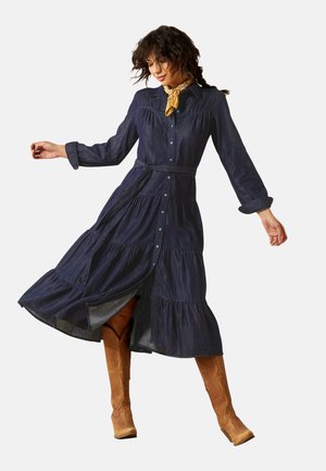 Frau in einem dunkelblauen, geknöpften Midikleid mit Gürtel, einem gelben Halstuch und braunen Cowboy-Stiefeln, die mit weit ausgestreckten Armen posiert.