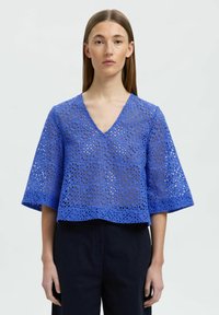 Blå, kort blus med V-ringning och vida ärmar. Tillverkad av textil med strukturerad yta som har cirkulära utskärningar, vilket skapar en ventilerande design.