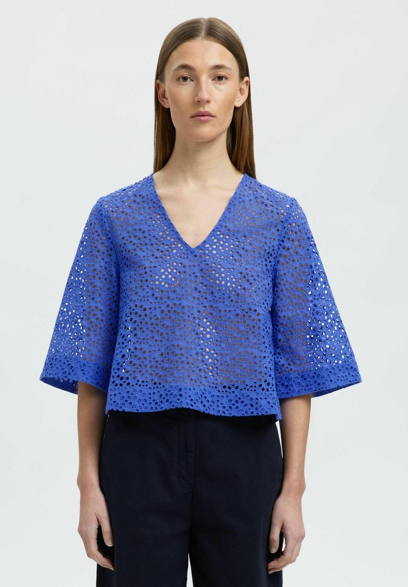 Blå, kort blus med V-ringning och vida ärmar. Tillverkad av textil med strukturerad yta som har cirkulära utskärningar, vilket skapar en ventilerande design.
