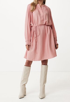 Frau trägt ein locker sitzendes, hellrosa Langarmkleid mit Taillengürtel, kombiniert mit kniehohen weißen Stiefeln mit Absatz, steht vor einem einfarbigen Hintergrund.