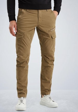 Man wearing brown cargo pants with flap pockets and white sneakers, standing with hands in pockets against a plain background.