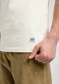 Cream cotton t-shirt with a square blue and white label at the hem, paired with brown corduroy pants featuring vertical ribbing.