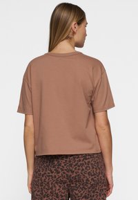 T-shirt in cotone marrone con una vestibilità rilassata, maniche corte e orlo leggermente accorciato, mostrato da dietro. Nessun motivo o accessorio distintivo.