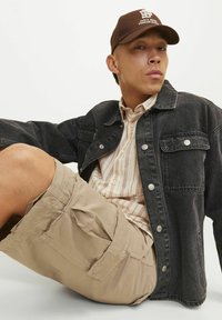 Sort denimjakke, beige stribet skjorte og beige cargo shorts. Brun kasket med logo. Casual pasform med shorts med lommer og knapdetaljer.
