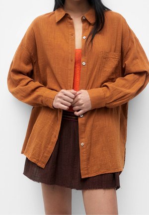 Person mit langen dunklen Haaren, die ein übergroßes, gebrannt oranges Hemd über einem orangen Top und einem braunen, strukturierten Rock trägt, Hände verschränkt.