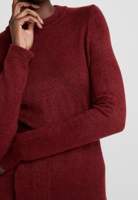 Maglione a maglia borgogna con collo rotondo e maniche lunghe. Presenta una texture morbida e polsini a coste, offrendo una vestibilità rilassata.