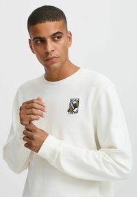 Cream-colored sweatshirt with a round neck, ribbed cuffs, and a small embroidered "Born Lucky" patch featuring a duck. Soft texture.