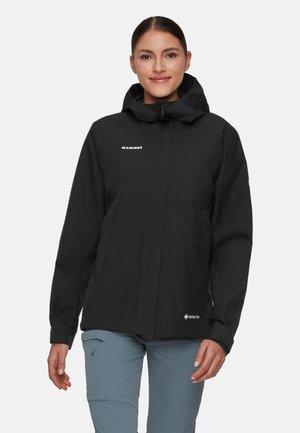 Kobieta ubrana w czarną kurtkę z kapturem Mammut GORE-TEX i niebieskie spodnie, stojąca i uśmiechająca się na jednolitym jasnym tle.