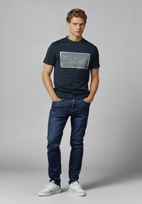 Marineblauwe katoenen t-shirt met een grijze rechthoekige "RED BULL RACING" grafiek, gecombineerd met donkere jeans en witte sneakers.