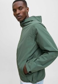 Grønn vanntett jakke med hette, justerbare mansjetter og sidelommer. Har en glatt, blank tekstur og elastisk midje.