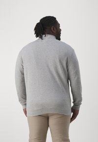 Sudadera gris claro de manga larga y cuello alto, con una textura suave y un dobladillo ligeramente curvado. Llevada con pantalones beige.