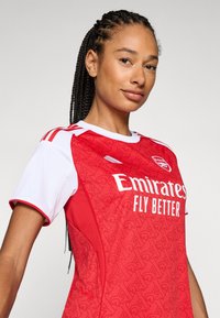 Maillot de football rouge avec des accents blancs, présentant un motif texturé, un col rond et le slogan "Emirates Fly Better" imprimé en blanc.