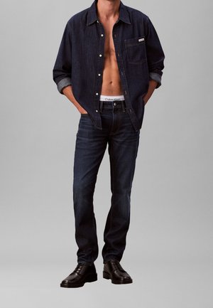Mann trägt ein aufgeknöpftes dunkles Jeanshemd, dunkle Jeans, schwarze Lederschuhe, mit sichtbarem Calvin Klein Bund unter dem Hemd.